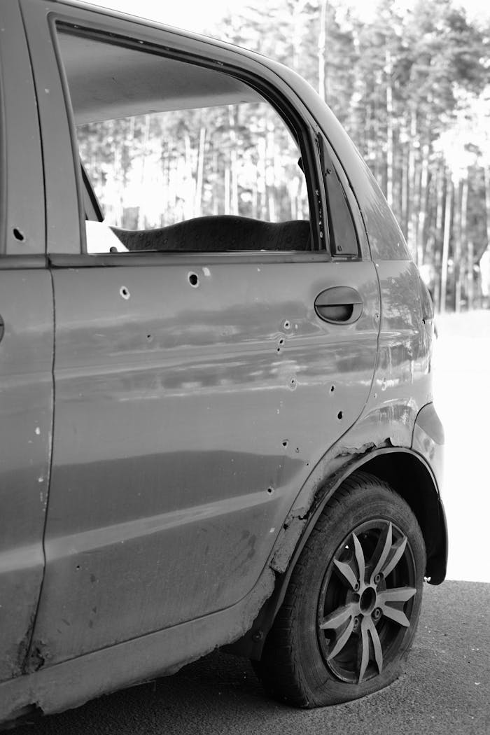 about-04 Close-up of a car door with bullet holes and a flat tire in a forest setting.